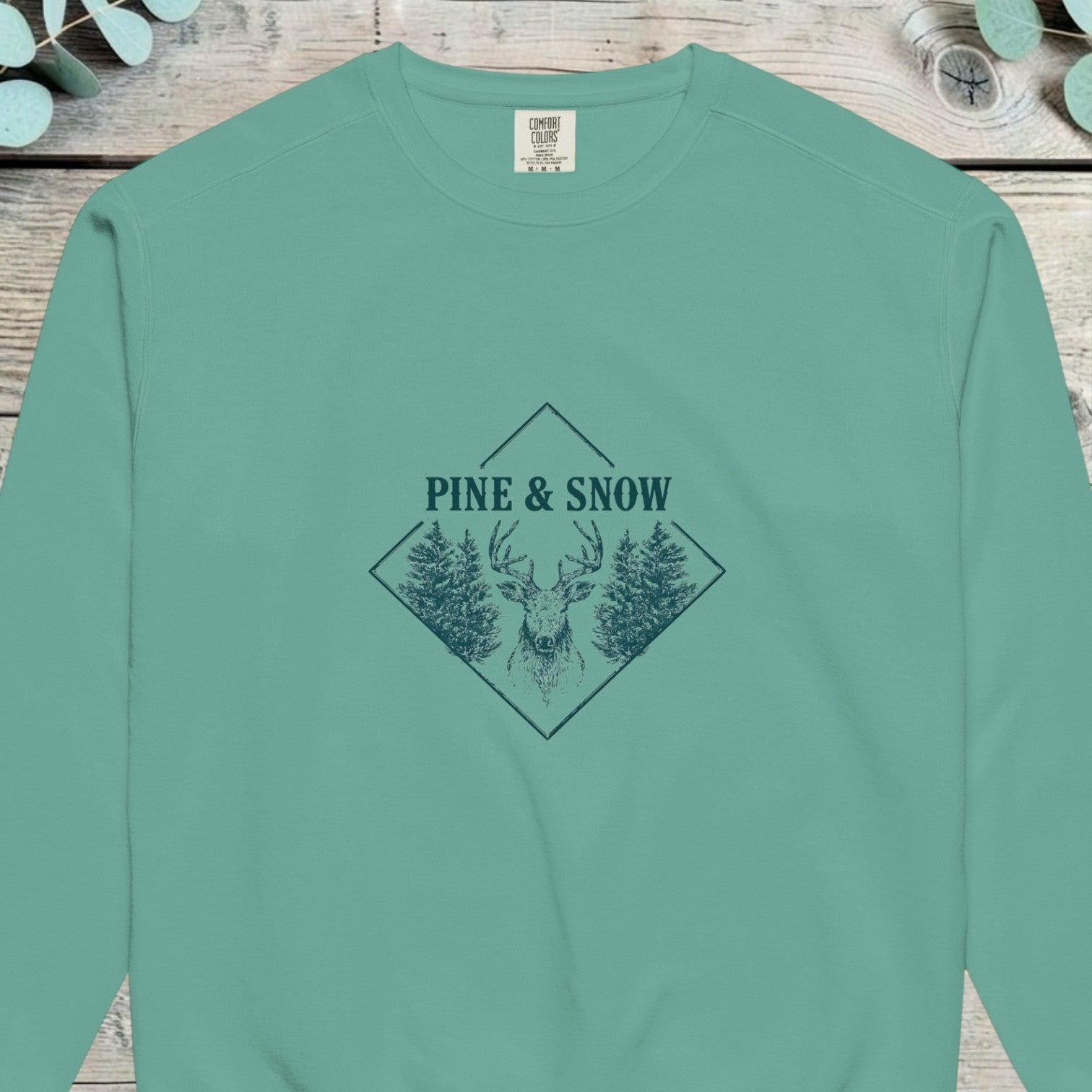 Stag head between pines in a diamond frame with PINE AND SNOW above, on a sweatshirt