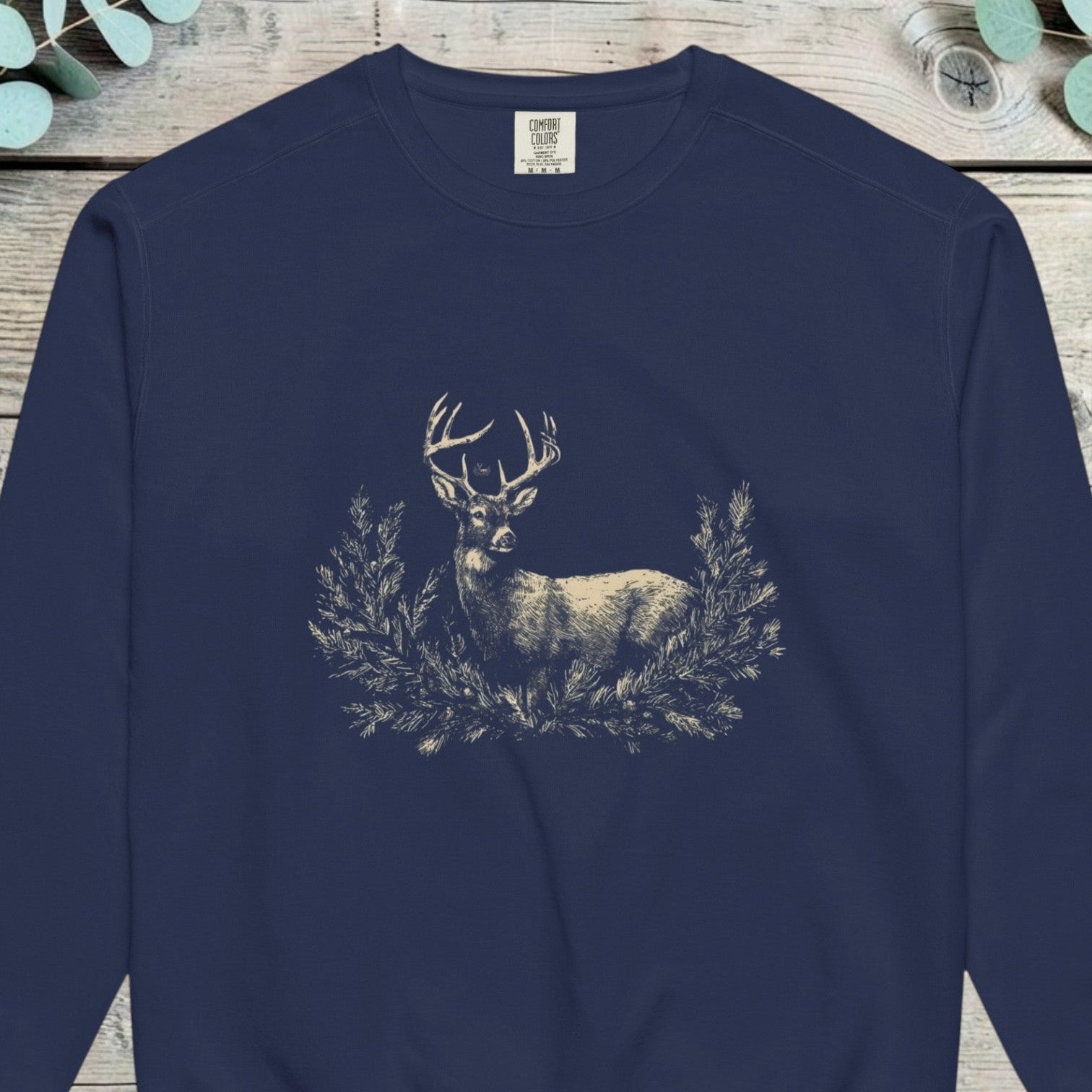 Pale stag standing among pine boughs on a deep navy sweatshirt